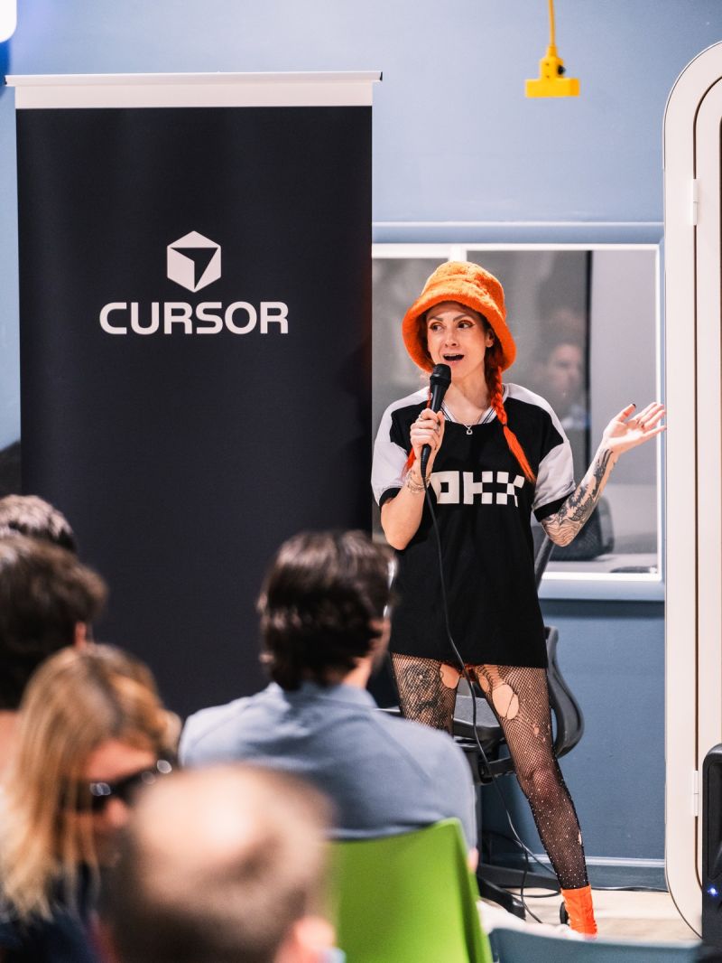 A speaker in an OKX shirt holding a microphone in front of a Cursor banner