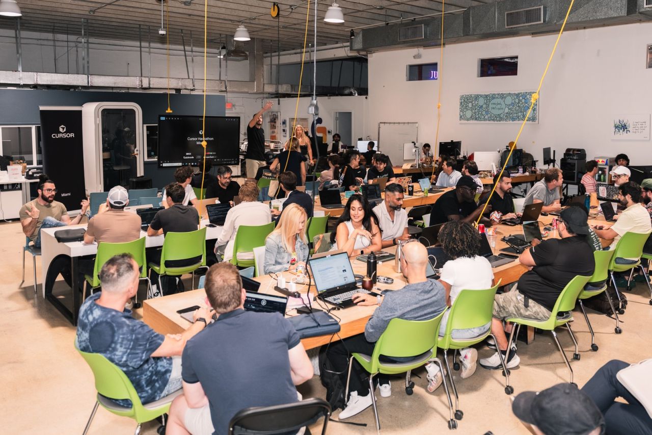 Wide view of the hackathon room at The LAB Miami, dozens of builders at long tables with laptops, a Cursor Hackathon Miami slide on screen